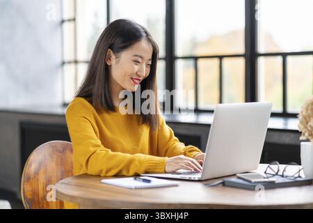 Asian woman working on laptop at home or in cafe. Young lady in bright yellow jumper sitting at desk typing on computer. Business oriental female in f Stock Photo
