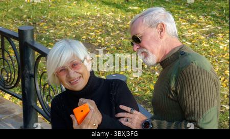Portrait of a loving elderly couple with a smartphone in hand on the background of the autumn city park Stock Photo