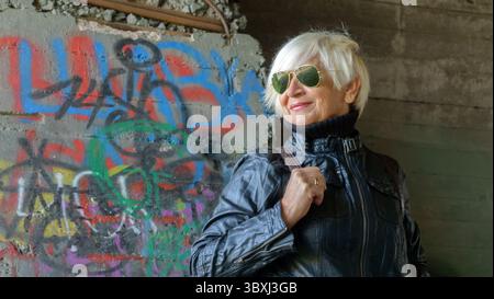 Portrait of a confident elderly woman in a black leather jacket wearing sunglasses against the background of old concrete walls Stock Photo