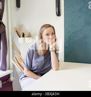 Business woman sitting at table and smiling in ceramic workshop. Young businesswoman at pottery workplace. Cheerful young female in blue dress looking Stock Photo