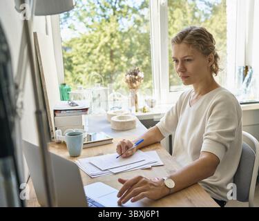 Work from home. Young female entrepreneur designer writing on paper sheet, looking at laptop, serious woman thinking and planning, happy lady in sunny Stock Photo