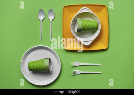 Colorful plastic disposable cutlery, such as two plates, spoons, forks, folded cups and one bowl on green background with copy space. The concept of p Stock Photo