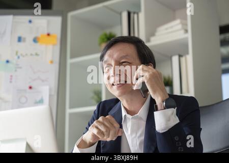 An elderly businessman smiling while talking on the phone, engaged in a discussion about data insights in a contemporary office filled with charts and Stock Photo