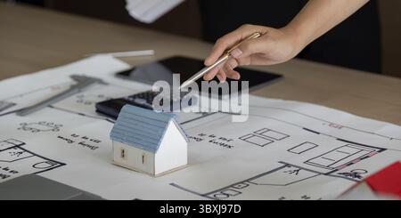 A close-up of a hand discussing a small house model on top of blueprints. This exchange highlights the essential planning that goes into construction Stock Photo