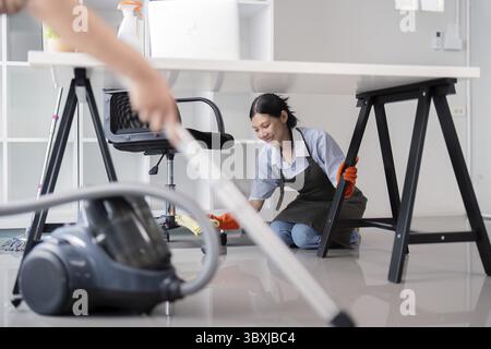 Female office cleaner kneeling and wiping the floor under a desk with a vacuum cleaner in the foreground, emphasizing thorough workplace cleaning Stock Photo