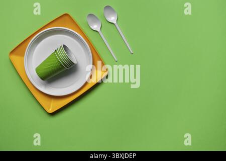Colorful plastic disposable tableware, such as two plates and spoons, folded cups on green background with copy space. The concept of picnic utensil. Stock Photo