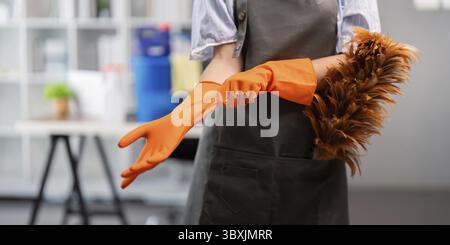 Female office cleaner putting on orange gloves while holding a duster, emphasizing workplace cleanliness Stock Photo