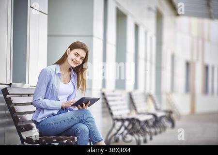 Ein junges Maedchen sitzt auf einer Bank, haelt ein Tablet in der Hand und laechelt Stock Photo