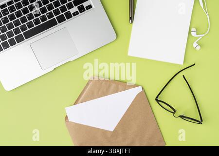 Blank paper sheet in craft envelope on green background. Metallic laptop keyboard, empty notebook, white earphones, glasses, pen. Horizontal from abov Stock Photo