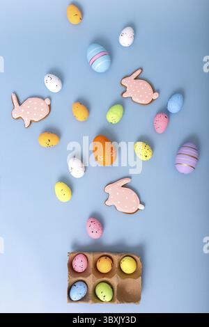 Beautiful decorative multicolored eggs and pink gingerbread in the form of rabbit, scattered on a light blue background, top view. Vertical Stock Photo