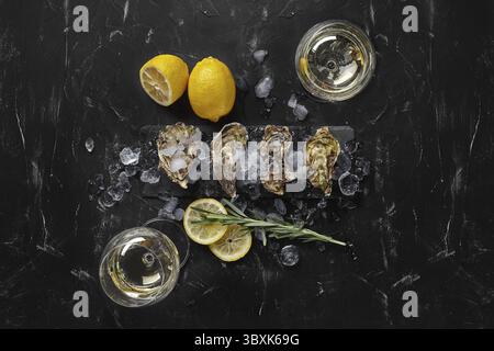 Fresh tasty closed oysters, ice, lemon and a green twig of lavender on a rectangle slate and two glasses of champagne are on a black stone textured ba Stock Photo