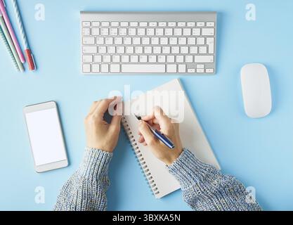 Faceless woman writing in notepad. Mobile phone with blank white screen display. Bright blue modern desk with smartphone. Top view. Copy space, mock u Stock Photo