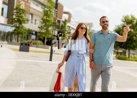 A couple walks hand in hand, dressed casually, absorbing the warm sunlight. They carry shopping bags while exploring a modern urban area filled with g Stock Photo