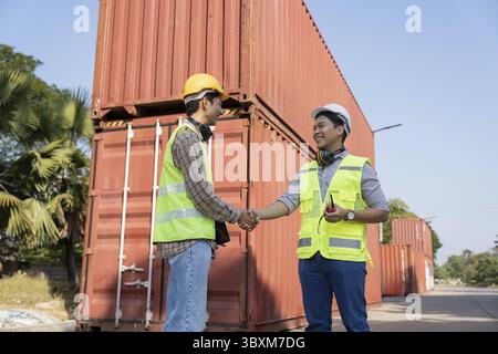 Two engineers in safety vests and helmets shake hands in front of shipping containers, symbolizing collaboration in logistics and industrial managemen Stock Photo