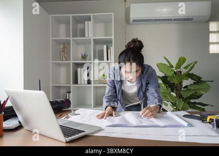 A focused engineer analyzes blueprints at her desk, surrounded by office supplies and a laptop, in a modern office environment Stock Photo
