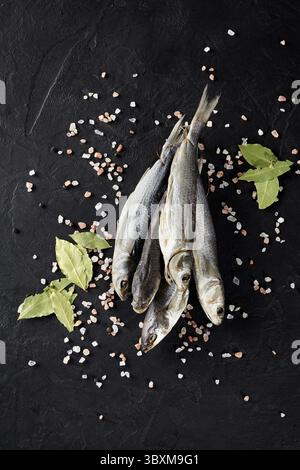 Bunch of dried mullet lying on black textured surface with scattered crystals of coarse Himalayan salt, peppercorns and dry bay leaves Stock Photo