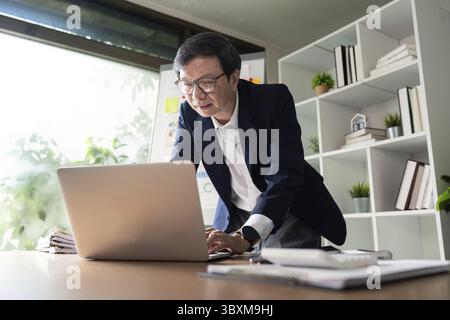 An elderly businessman focused on his laptop, analyzing data and preparing for financial management, surrounded by documents and a modern office envir Stock Photo
