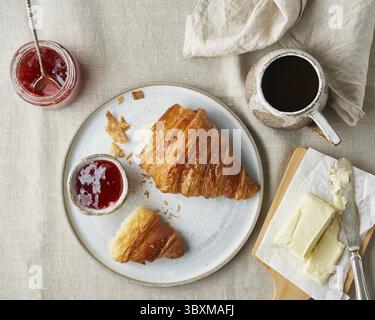 One delicious croissants on plate and hot drink in mug. Morning French breakfast with fresh pastries and jam. Light gray background, top view Stock Photo