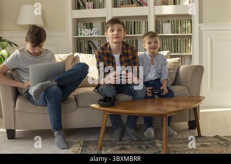 Three brother boys are sitting at home on a sofa in the sun playing computer games on a laptop and video games Stock Photo