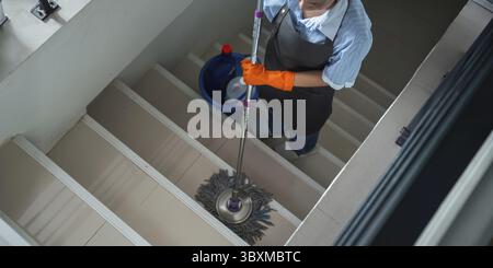 A cleaning woman is standing inside a building holding with a blue tank on the side chemicals and facilities for tidying up in her hand Stock Photo