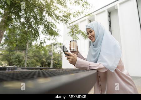 A joyful Muslim woman in a hijab smiles as she uses her smartphone while holding a coffee cup, embodying a modern and connected lifestyle in a busines Stock Photo