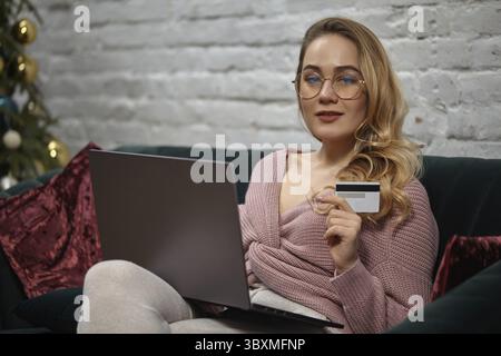 Female blogger in pink cardigan, pants, glasses. Sitting on green sofa with burgundy pillows, showing plastic card, holding laptop and smiling, posing Stock Photo
