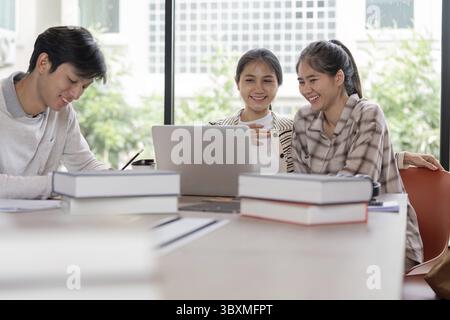 A group of cheerful students collaborating on a project with laptops and books in a bright, modern classroom setting Stock Photo
