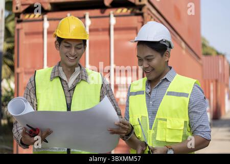 Two professionals in safety vests smile while examining blueprints, showcasing teamwork and planning in logistics and engineering on-site Stock Photo