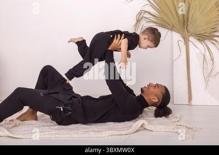 Stylish father and son, in black clothes, lying on a knitted blanket, on the floor in the room, having fun playing. The father lifts his son above him Stock Photo