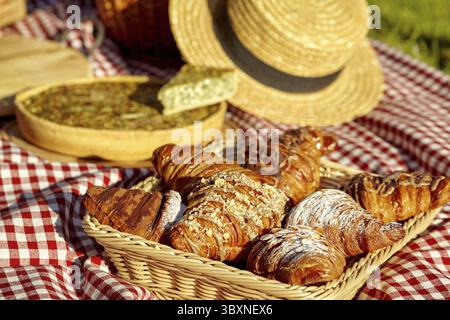 Wicker tray with sweet fluffy puff croissants on red and white checkered blanket on blurred background with savory quiche and straw hat during cozy pi Stock Photo