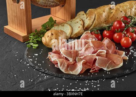Jamon manually carved from pig leg on a jamonera stand, cherry tomatoes, baguette, herbs, scattered spices on black stone slate board. Dark grey backg Stock Photo
