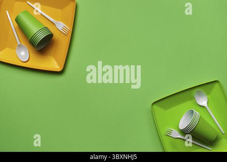 Colorful plastic disposable tableware, such as two plates, spoons, forks, folded cups on green background with copy space. The concept of picnic utens Stock Photo