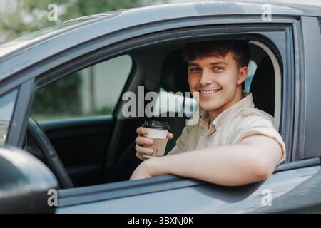 Portrait of handsome young man holding paper cup of fresh coffee while sitting inside his car. Young male smiling and looking at camera. Coffee to go. Stock Photo