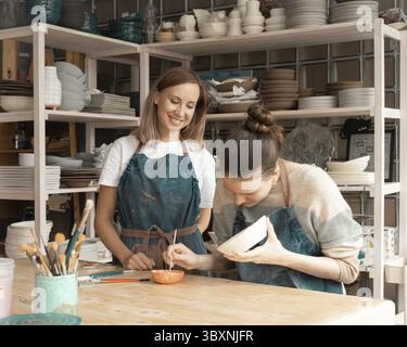 Woman making ceramic pottery. Attractive skilled young lady in apron standing at table and teaching student. Concept for workshop and master class Stock Photo