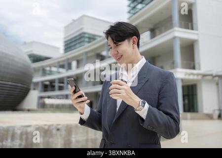 A young professional man in a suit smiles while using his smartphone and holding a coffee cup outside a modern office building Stock Photo