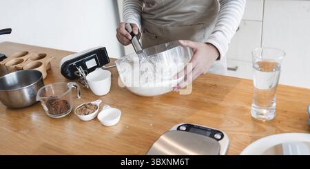 A cozy home baking kitchen with a person mixing batter in a glass bowl, surrounded by various baking ingredients and utensils on a wooden countertop Stock Photo