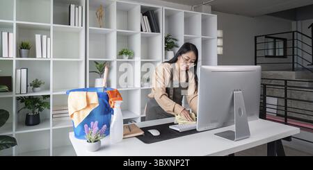 Housekeeper in uniform cleaning a modern office desk with computer and supplies, ensuring a tidy and organized workspace Stock Photo