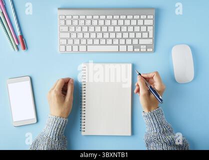 Faceless woman writing in notepad. Blank white notepad. Mobile phone with blank white screen display. Bright blue modern desk with smartphone. Top vie Stock Photo