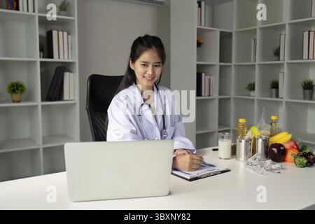 The nutrition expert is reviewing health-related data on a laptop while taking notes about nutritious food options arranged on the table, showcasing a Stock Photo