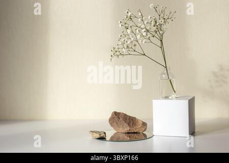One branch of a gypsophila flower standing on a white paper cube, near three stones on a round mirror, on a beige background, copy space, abstract des Stock Photo