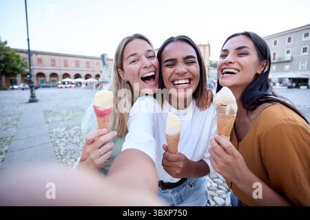 Three happy female friends taking a selfie while eating ice cream in a town square Stock Photo