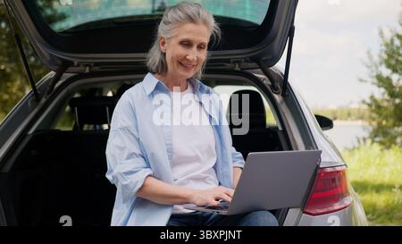 Happy positive Caucasian mature elderly business woman senior female freelancer aged businesswoman worker tourist sit car auto trunk working laptop Stock Photo