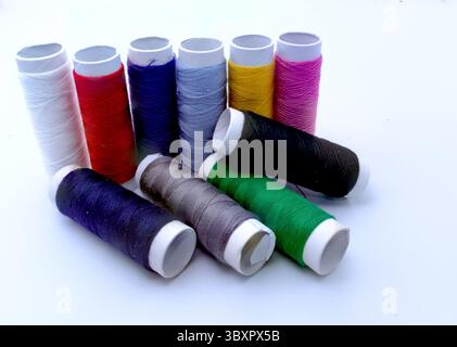 Ten spools of vibrant sewing thread in white, red, blue, light blue, yellow, pink, black, brown, purple, and green are arranged on a white surface. Stock Photo
