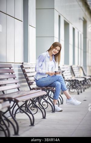 Ein junges Maedchen sitzt auf einer Bank, haelt ein Tablet in der Hand und laechelt Stock Photo