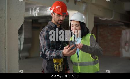 Two multiethnic builders using mobile phone building design app showing thumb up Japanese woman Caucasian man hardhat engineers business inspector Stock Photo