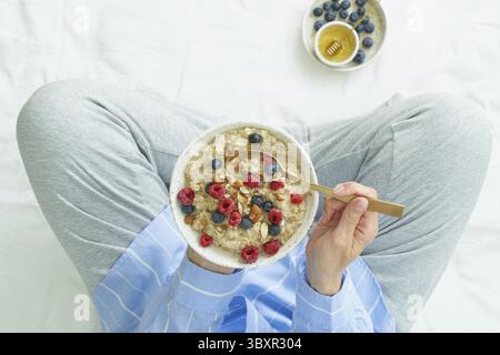 Top view faceless woman holding plate of oatmeal, sitting in bed early morning, quiet home environment. Healthy breakfast Stock Photo
