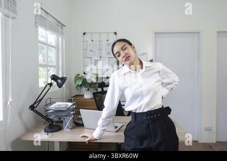 A young professional woman in a modern office setting experiencing back pain, leaning against her desk with a pained expression Stock Photo