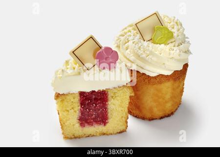 Appetizing whole and sliced fluffy vanilla cupcakes with raspberry filling topped with swirls of whipped cream sprinkled with coconut shavings and cho Stock Photo