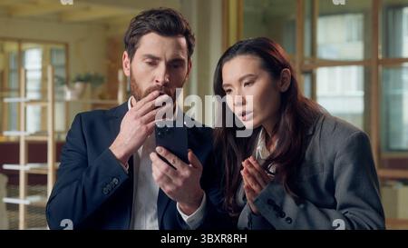 Thoughtful worried two business man woman look mobile phone read bad news reject at office Caucasian Korean businesswoman businessman anxious confused Stock Photo