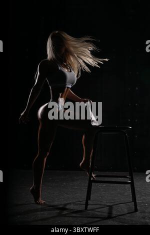 blonde woman athlete confidently poses on stool, displaying her strength and athleticism in dramatic dark studio setting during professional photoshoot Stock Photo